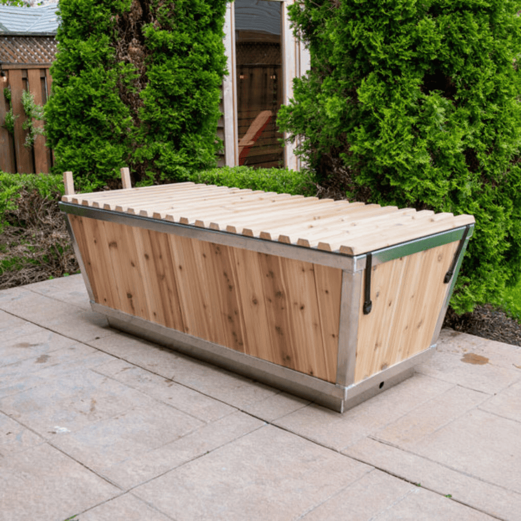 Corner view of Polar cold plunge with wood roll-top cover, placed on concrete with pine shrubs in the background